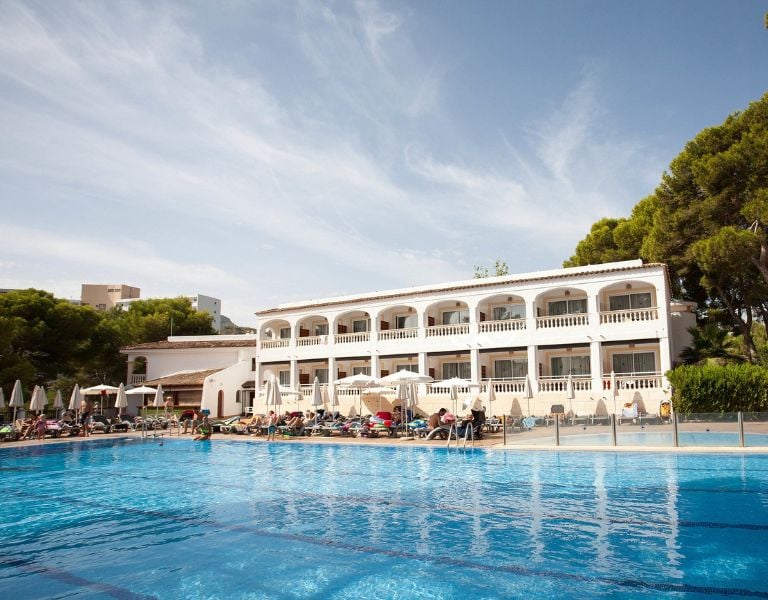 Outdoor pool at a beach club, with a Mediterranean-style building, surrounded by sunbathers and pine trees.
