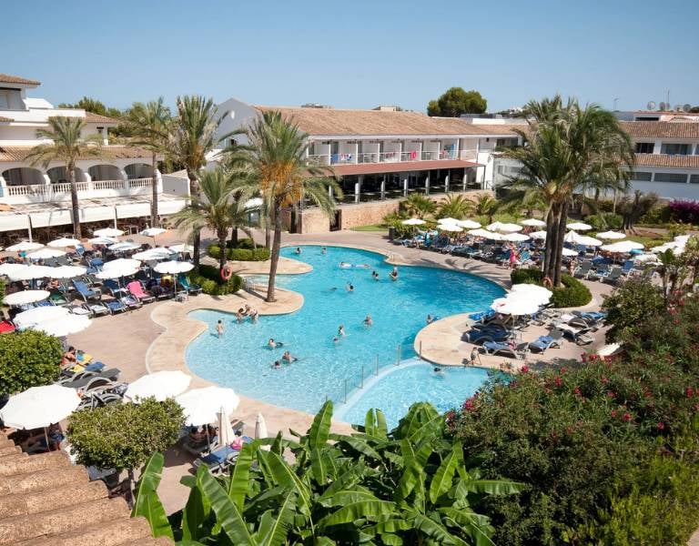Aerial view of Beach Club Es Talaial with a large swimming pool, sun loungers, and tropical palm trees.