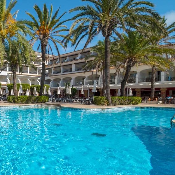 Swimming pool at a beach club with clear blue water, surrounded by sun loungers and palm trees, hotel building in the background.