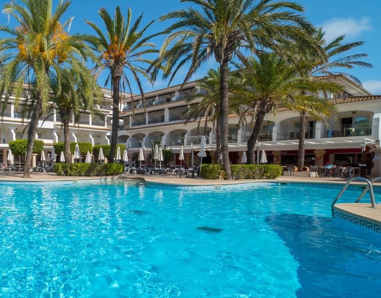 Swimming pool at a beach club with clear blue water, surrounded by sun loungers and palm trees, hotel building in the background.