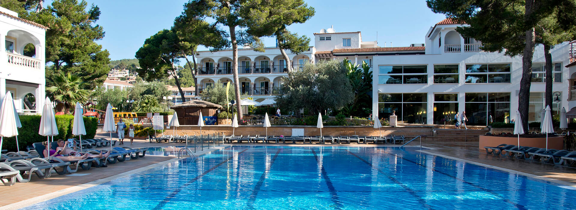 Outdoor pool at beach club with sun loungers, parasols, and a Mediterranean-style building surrounded by trees.