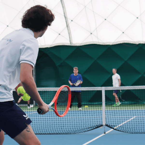 Tennis coach instructing adult learners on an indoor court, with players holding rackets and practising skills.