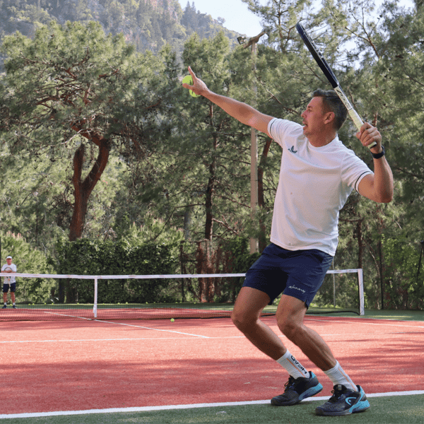 Tennis coach serving on a red clay court with students in the background, surrounded by trees and greenery.