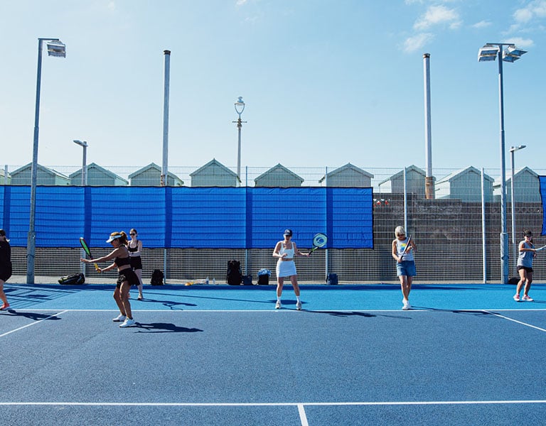 Outdoor tennis match in Brighton with players on a blue court, seaside beach huts in the background under a clear sky.