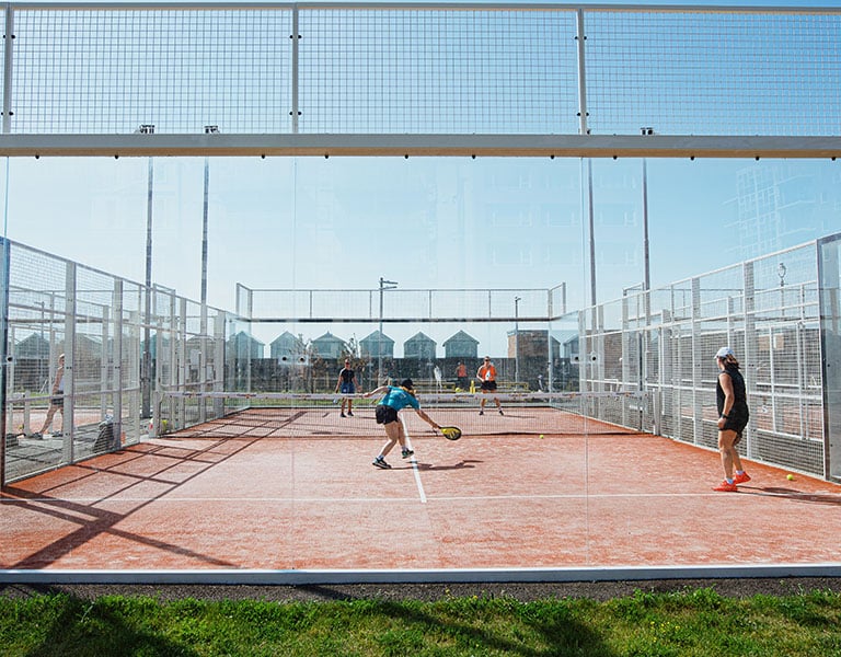 People playing padel tennis on a fenced outdoor court in Brighton with blue skies overhead and modern houses in the background.