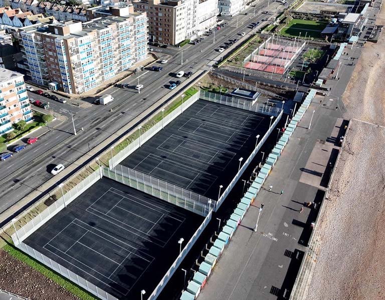 Aerial view of tennis and padel courts near the seafront in Brighton, with roads, buildings, and the beach in the background.