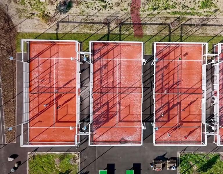 Aerial view of three orange padel courts with players, surrounded by greenery, during a tennis padel weekend in Brighton.