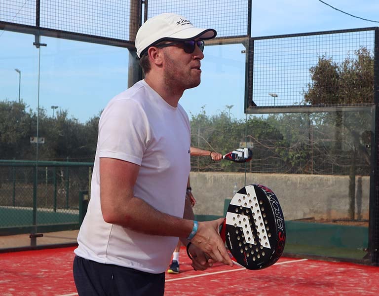 Man wearing a white t-shirt and cap, holding a padel racquet on a court in Brighton, ready to play.