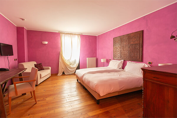 Hotel room with purple walls, twin beds, wooden floor, dresser, wall-mounted TV, sofa, and desk with chair at Hotel Cristallo.