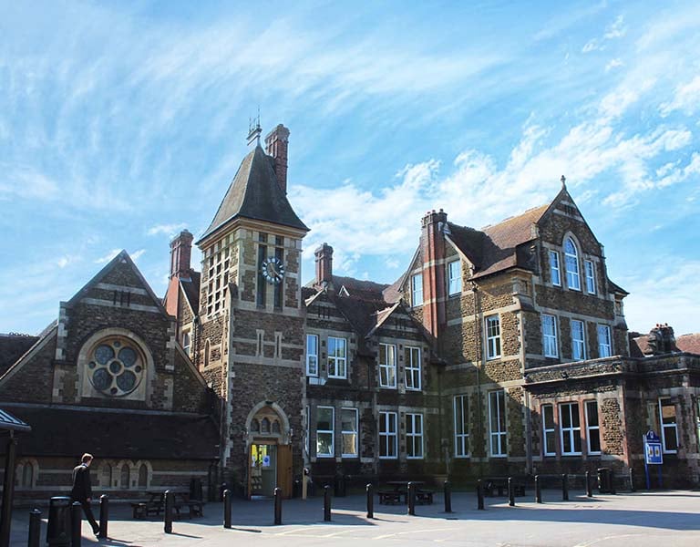 Wide shot of Churcher's College exterior, featuring a clock tower, arched windows, and decorative brickwork under a blue sky.