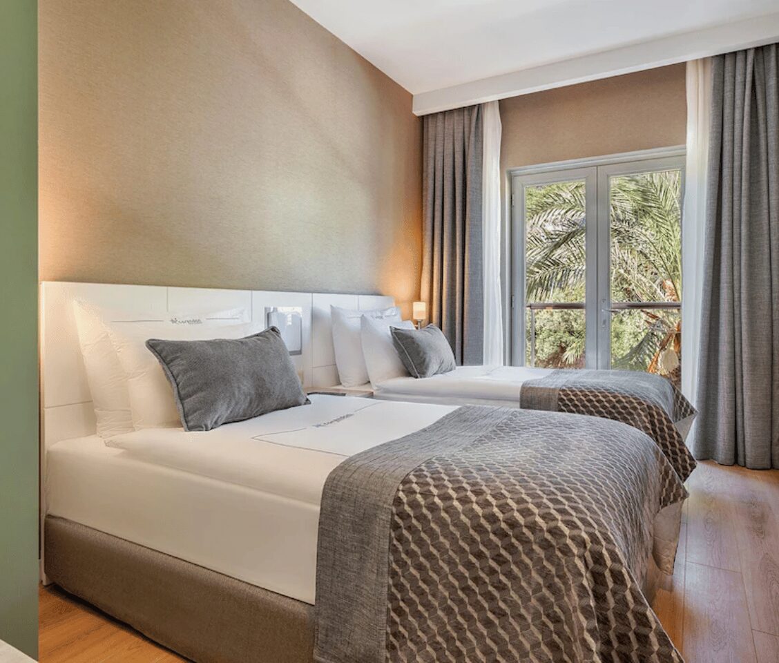 Corendon family room with two double beds, elegant bedding, modern decor, and a view of greenery through a large window.