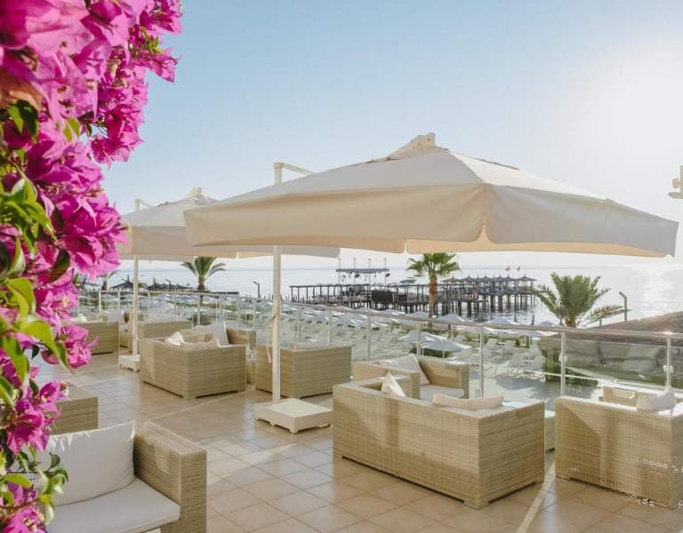 Corendon Gallery beachfront lounge with wicker seating, white umbrellas, pink bougainvillaeas, ocean view, and a wooden pier.