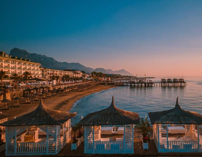 Beachfront view at sunset with luxurious straw-roofed cabanas, distant mountains, calm sea, sunbeds, and a grand hotel.