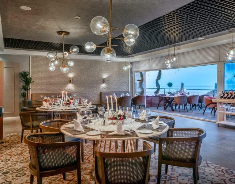 Elegant dining room at Corendon Gallery hotel with round tables, white tablecloths, and modern hanging lights, overlooking a scenic view through large windows.