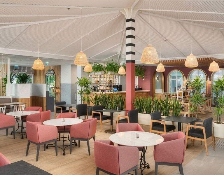 Indoor dining area at Corendon Hotel with pink chairs, round tables, potted plants, and wicker pendant lights under a vaulted ceiling.
