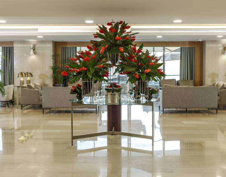 Luxurious hotel lobby with red flowers on a glass table, grey sofas, and large windows.