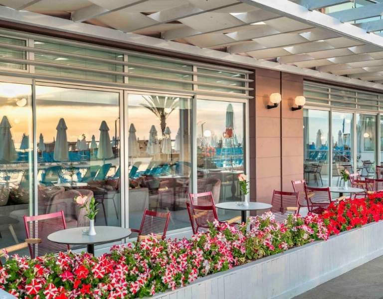 Outdoor dining area at Corendon hotel with red chairs, flower planters, and views of sun umbrellas and palm trees at sunset.