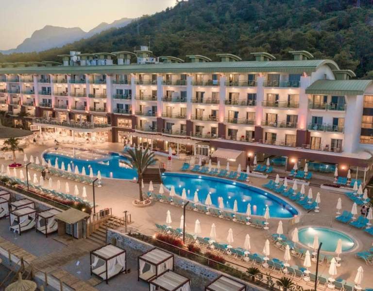 Aerial view of Corendon resort with a large swimming pool, sun loungers, umbrellas, and a hotel building set against green hills.