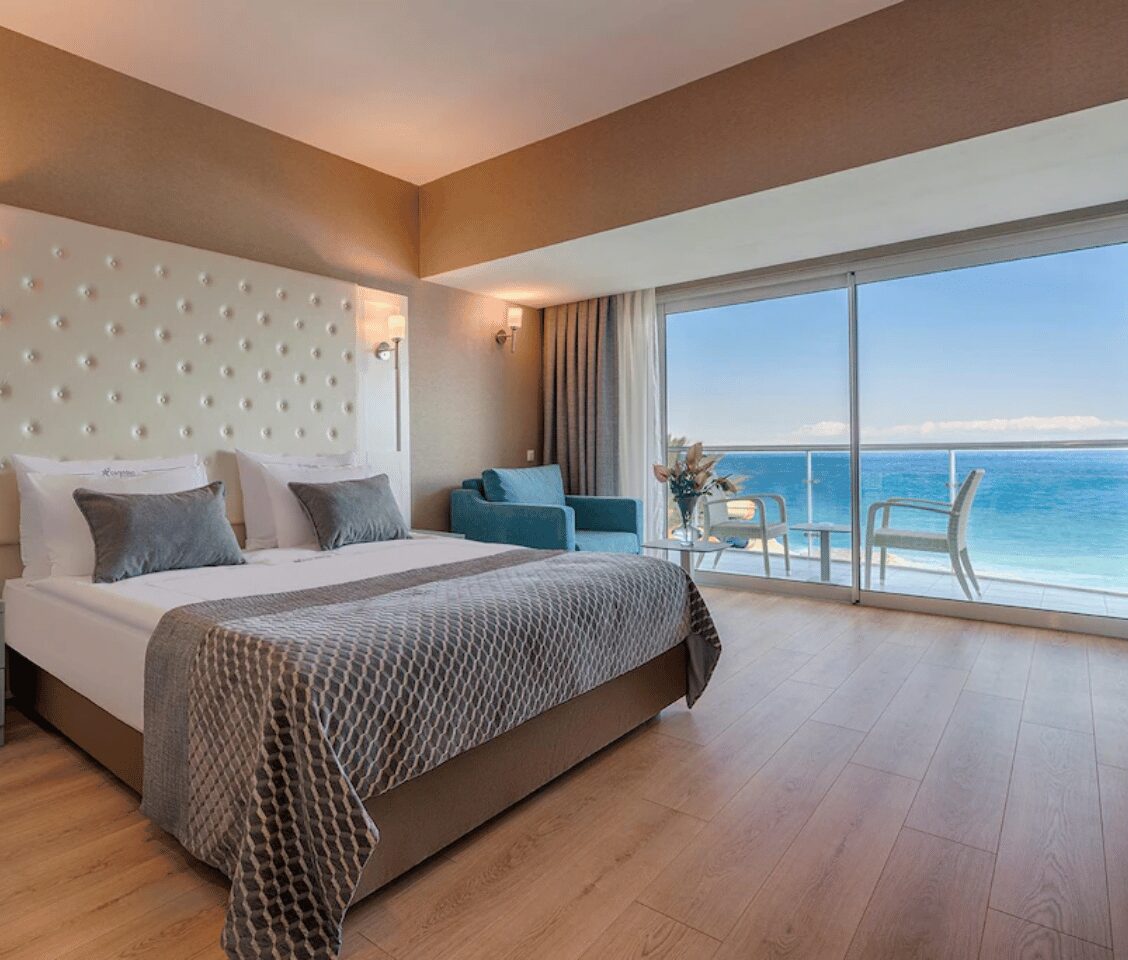 Luxurious hotel room with sea view at Corendon, featuring a double bed, beige headboard, armchair, balcony, and ocean scenery.