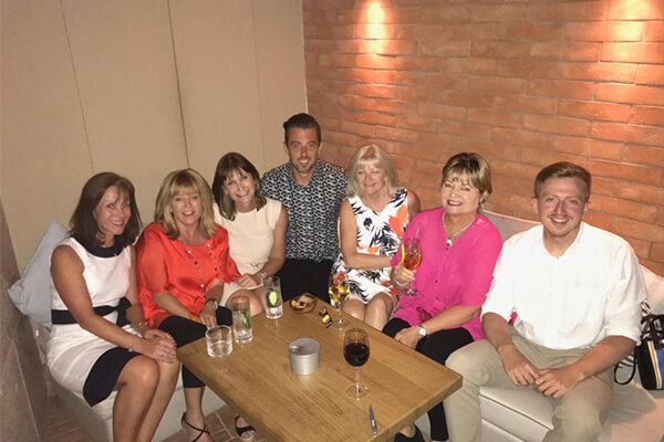 Group of six people sitting at a small table with drinks, smiling and enjoying themselves at Costa Navarino with a brick wall behind them.