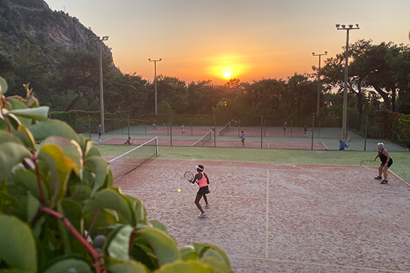 Tennis match at sunset on a clay court with surrounding greenery and floodlights, players visible on the court.