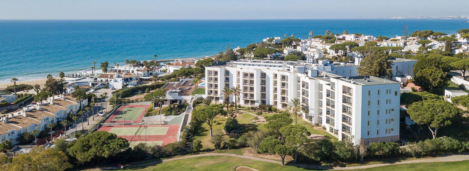 Aerial view of Dona Filipa Hotel in Algarve, Portugal, displaying modern white buildings, tennis courts, lush greenery, and nearby sandy beach.