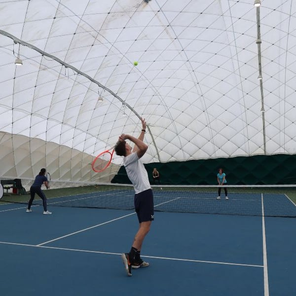 Indoor tennis coaching session in Beaconsfield, with a coach demonstrating a serve to players on the court.