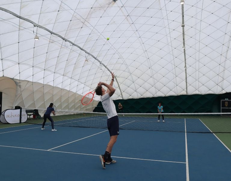 Indoor tennis coaching session in Beaconsfield, with a coach demonstrating a serve to players on the court.