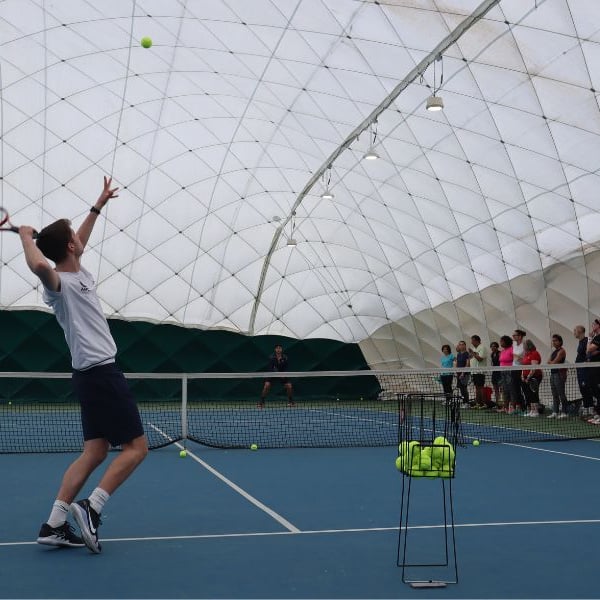 Tennis coach at DL Beaconsfield demonstrating a serve on an indoor court while a group of players observe.