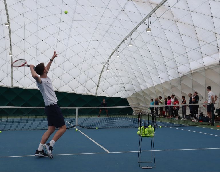 Tennis coach at DL Beaconsfield demonstrating a serve on an indoor court while a group of players observe.