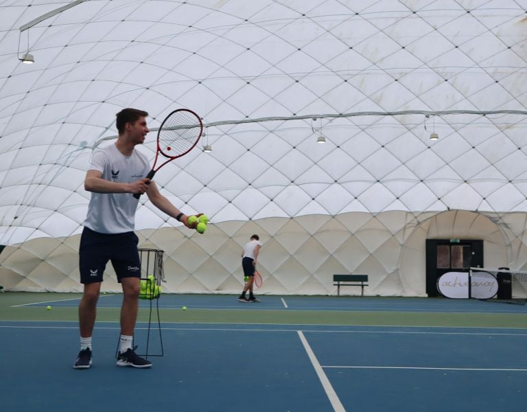 Tennis coaches on court under a dome, feeding tennis balls, with Active Away logo visible, indoors at Beaconsfield.