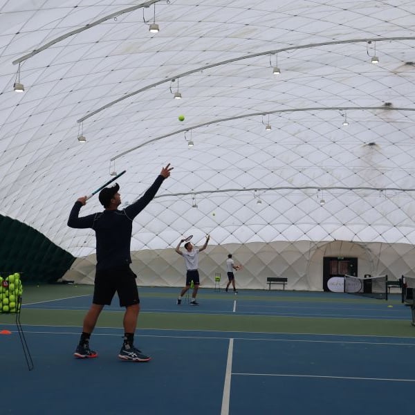 Tennis coaches serving on indoor bubble courts at David Lloyd Beaconsfield, with a basket of tennis balls and court equipment.