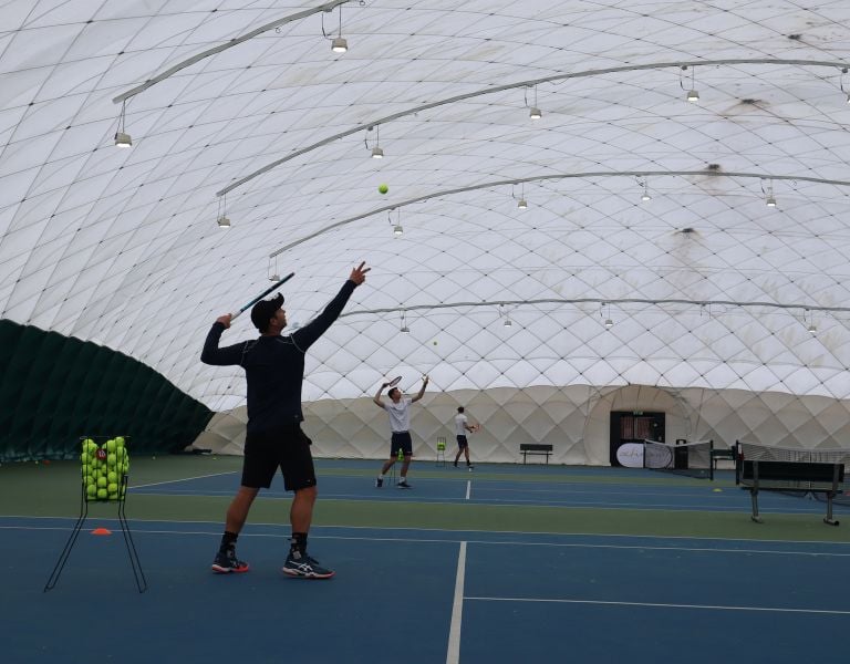 Tennis coaches serving on indoor bubble courts at David Lloyd Beaconsfield, with a basket of tennis balls and court equipment.