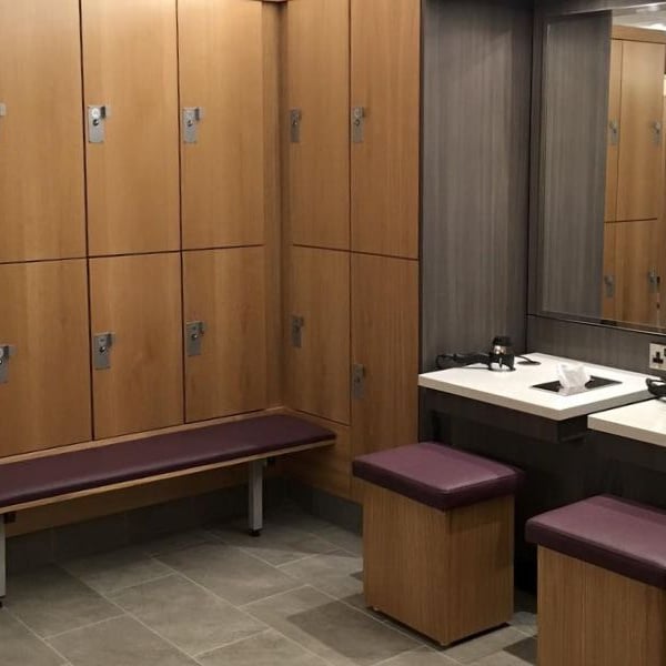 Modern changing room at DL-Beaconsfield with wooden lockers, upholstered seating, and a vanity area with mirrors and basins.