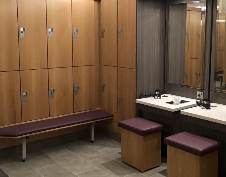 Modern changing room at DL-Beaconsfield with wooden lockers, upholstered seating, and a vanity area with mirrors and basins.