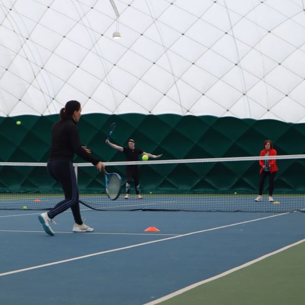 Three people on an indoor tennis court at David Lloyd Beaconsfield performing tennis drills; one player about to hit a ball with forehand stroke, two others standing near the net.