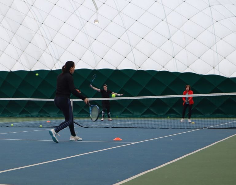 Three people on an indoor tennis court at David Lloyd Beaconsfield performing tennis drills; one player about to hit a ball with forehand stroke, two others standing near the net.