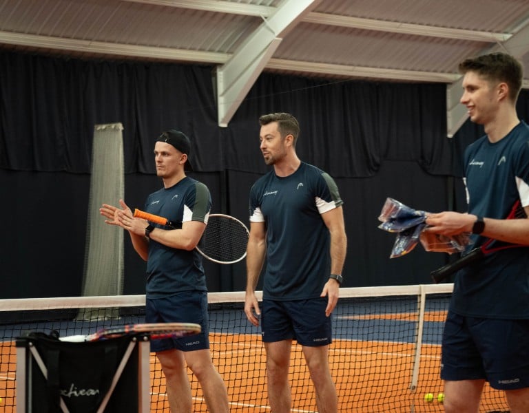 Three tennis coaches on an indoor court at David Lloyd Cheshire Oaks, wearing navy sports outfits and holding racquets.