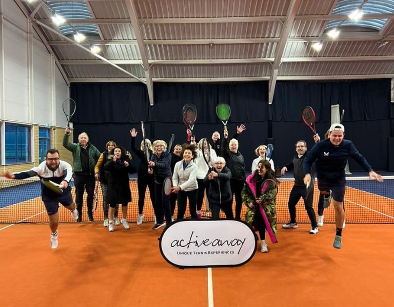 Group of enthusiastic tennis players posing on an indoor court at DL Cheshire Oaks, holding rackets behind an 'Active Away' banner.