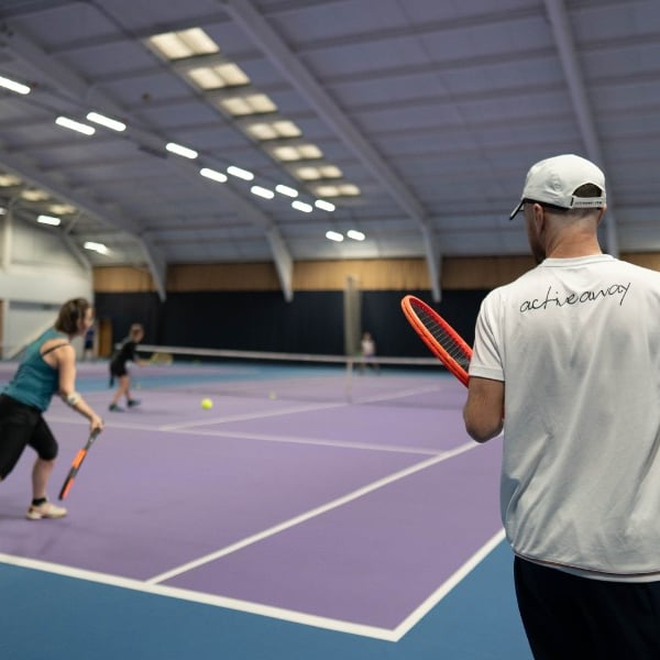 Tennis coach observes two players practising on a purple indoor court at DL Chorley, with vivid lighting and spacious layout.
