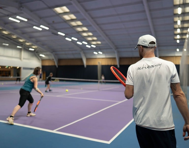 Tennis coach observes two players practising on a purple indoor court at DL Chorley, with vivid lighting and spacious layout.