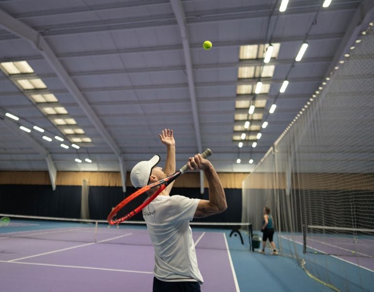 Tennis coach in a white cap serving on a purple indoor court, with bright overhead lights and another player seen in the background.