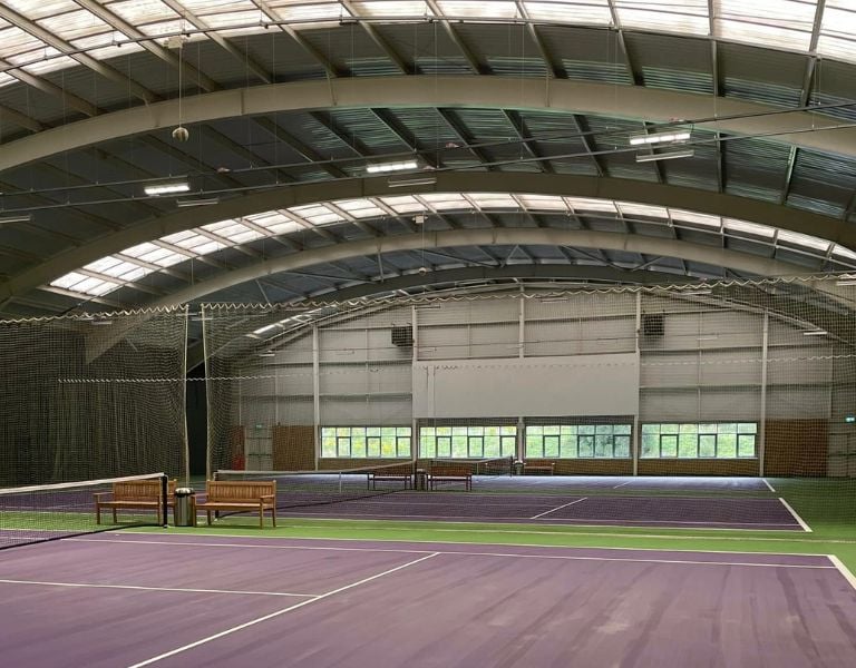 Indoor tennis court at DL Edinburgh with purple and green surface, large roof, natural light, and wooden benches.