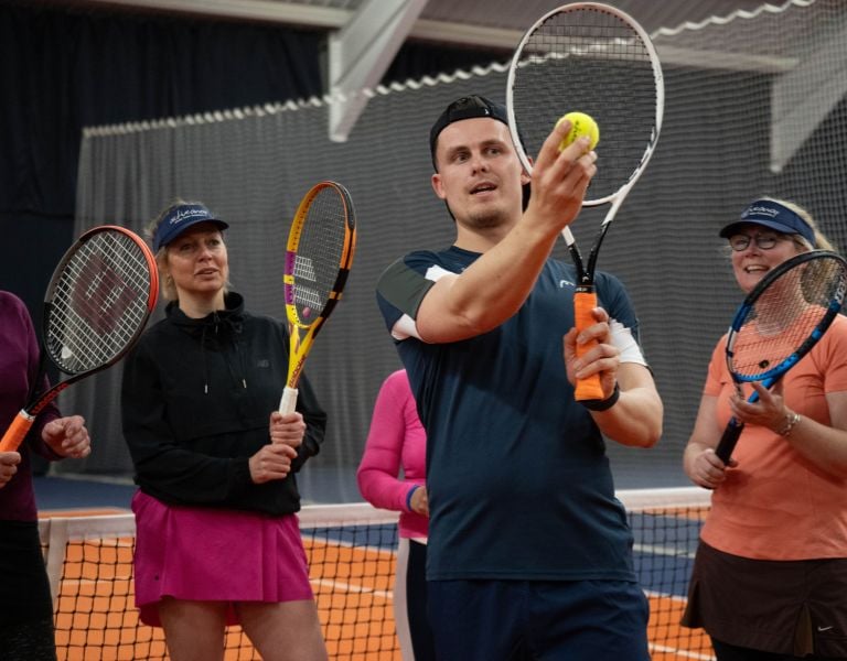 Indoor tennis coaching session with coach Alex showing a technique to adult players standing on a clay court.