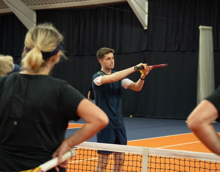 Tennis coach Jack demonstrates a forehand stroke to adult players at an indoor court in MK Gallery.