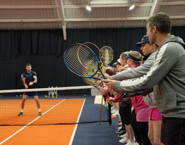Group of people during a tennis coaching session with instructor Josh at DL MK Gallery, all holding racquets and standing on an indoor court.