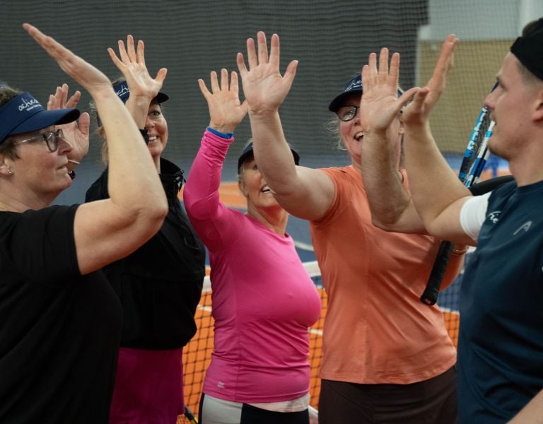 Five women in sports gear exchanging high fives on a tennis court, each holding a tennis racquet, showing teamwork and joy.
