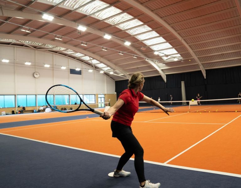 Woman playing tennis on an indoor orange court at David Lloyd Milton Keynes, preparing to return a shot with other players in the background.