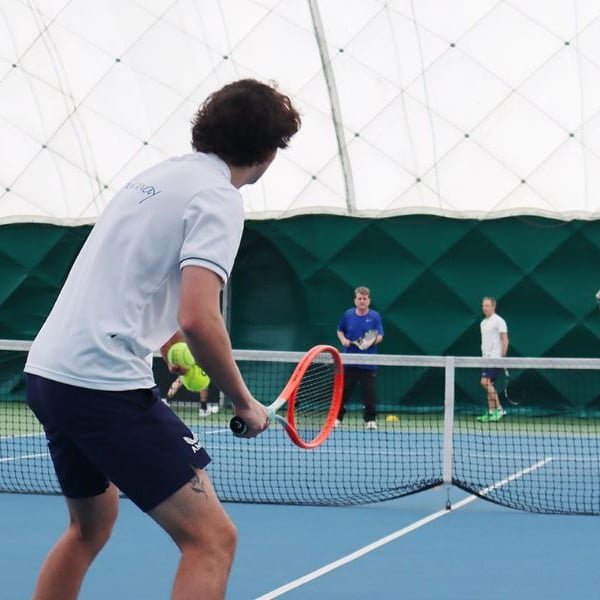 Group playing tennis indoors at DL Northwood Gallery, with a young man in white ready to hit the ball using an orange racquet.