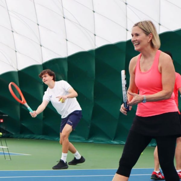 Indoor tennis lesson at DL Northwood with a coach smiling and a student preparing to hit a ball.